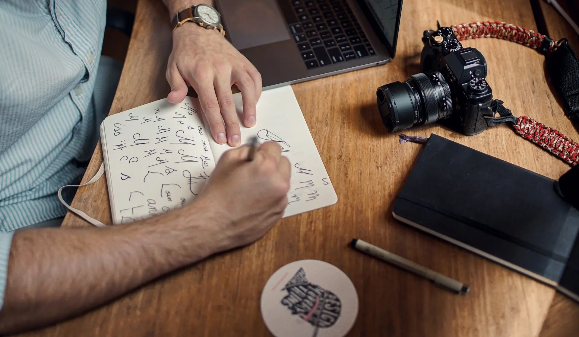 Une main dessine des lettres dans un carnet, un appareil photo posé sur le bureau.