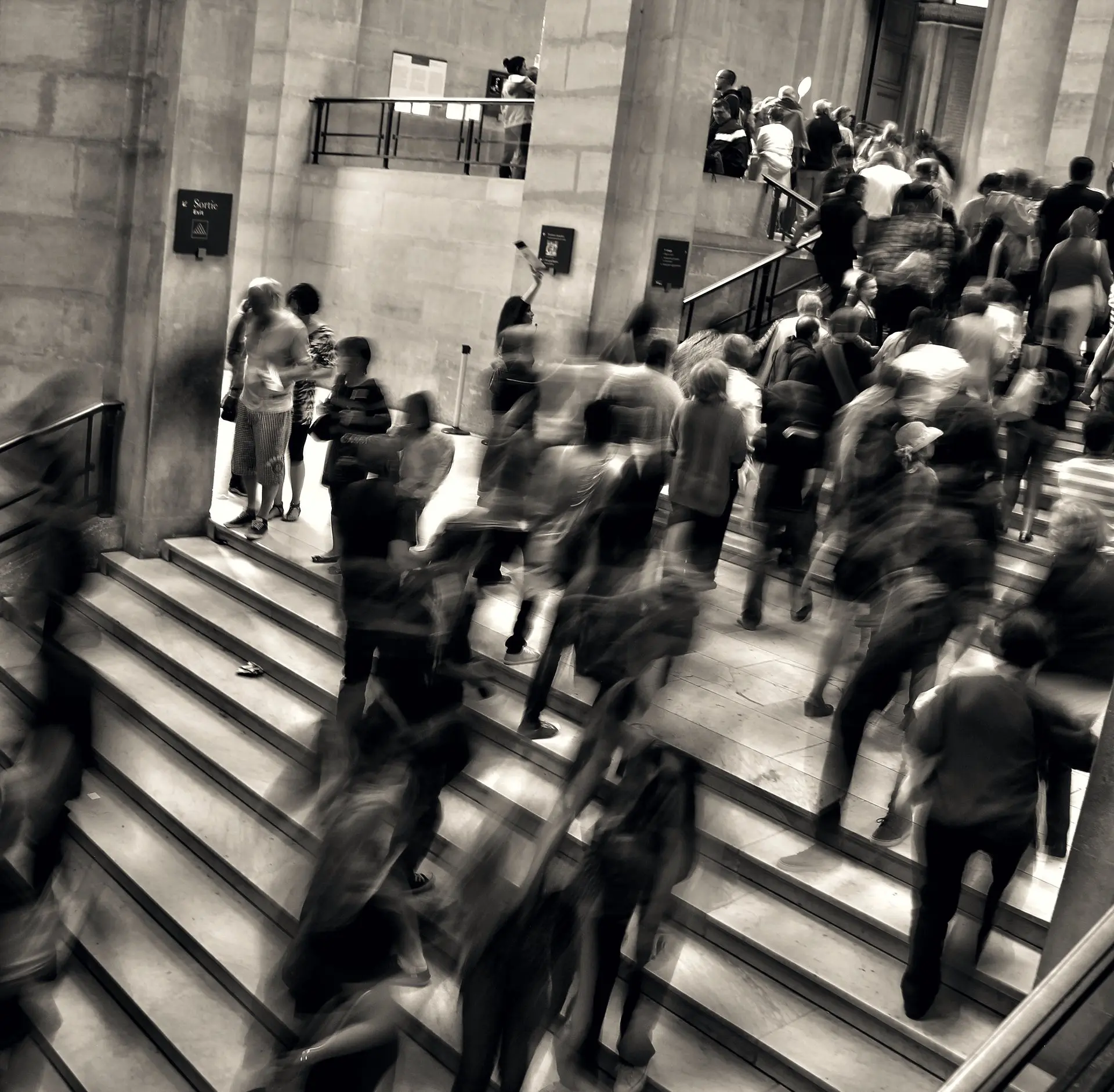 Une foule monte et descend l’escalier d’un grand hall, floutée par le mouvement.